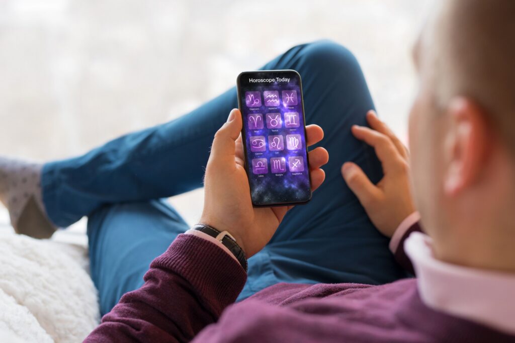 Horoscop3 - Tech World Muž pohodlne sedel na gauči, ktorý držal v ruke mobilný telefón. Displej spúšťa aplikácie s názvom